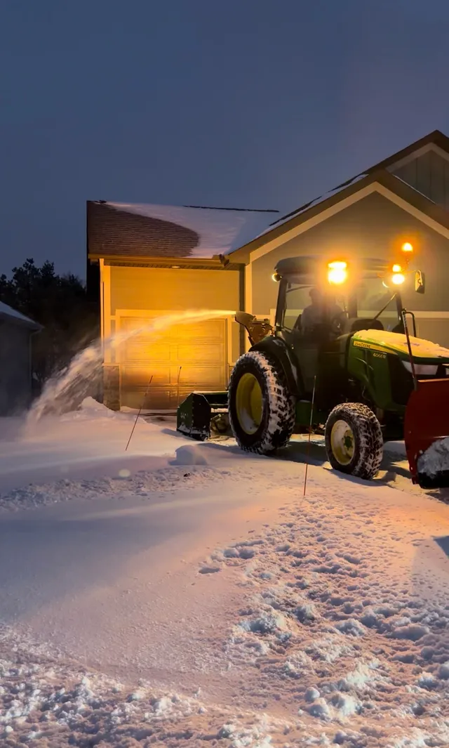 An Ecosnow tractor throwing snow clear of a driveway at night, work lights on and the garage lit up behind it.